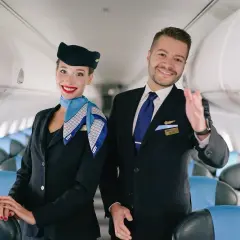 Uśmiechnięta stewardessa i steward w mundurach LOT stoją w samolocie, na tle rzędów siedzeń.