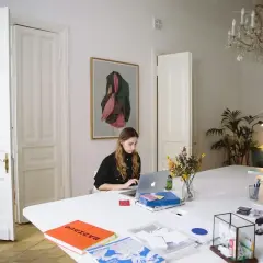 Kobieta pracuje przy laptopie w jasnym pomieszczeniu z obrazem na ścianie i bukietem kwiatów na stole.