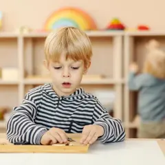 Chłopiec w bluzie w paski skupiony na zadaniu z materiałami Montessori. W tle inne dziecko przy półce.