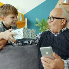 Chłopiec wręcza dziadkowi prezent. Dziadek, jako aktywny rodzic, zaskoczony trzyma telefon.