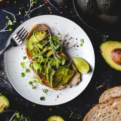 Tost z awokado, kiełkami i limonką na talerzu, obok kilka awokado i kromki chleba.