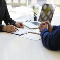 Dwie osoby przy biurku, jedna pisze na dokumencie, druga ma złożone ręce.