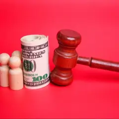 Drewniany młotek sędziowski, zwinięty banknot 100 dolarów i trzy drewniane figurki symbolizujące rodzinę. Pytanie: do kiedy alimenty na dziecko?