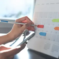 Dłoń z ołówkiem zaznacza datę na kalendarzu z kolorowymi karteczkami.