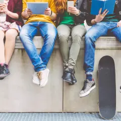 Cztery osoby siedzą na murku, każda z nich korzysta z telefonu, tabletu lub czyta książkę. Obok nich leży deskorolka.