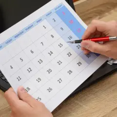 Dłoń z długopisem wskazuje na datę 10 na kalendarzu obok laptopa.
