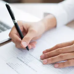 Dłoń pisze na dokumencie, obok laptopa. Ważne jest, ile jest ważne zaświadczenie o niekaralności przy wypełnianiu wniosków.