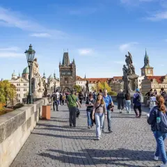 Most ludzi na moście Karola w Pradze, z widokiem na zabytkowe budynki i wieże.