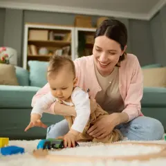 Uśmiechnięta mama bawi się z dzieckiem klockami i pociągiem na dywanie w salonie.