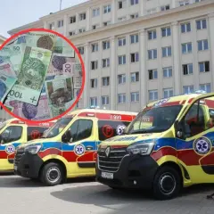 Polskie banknoty w czerwonym okręgu na tle kilku karetek pogotowia przed budynkiem.