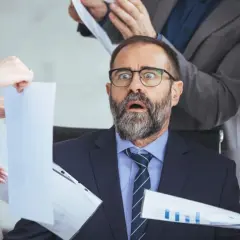Przerażony mężczyzna w garniturze otoczony przez ludzi z dokumentami.