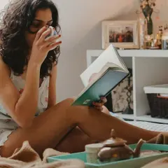 Kobieta w piżamie pije herbatę i czyta książkę w łóżku.
