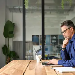 Mężczyzna w okularach pracuje na laptopie przy drewnianym biurku.