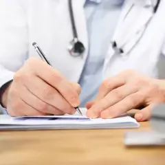 Lekarz w białym kitlu z długopisem pisze na dokumentach. Obok pieczątki.