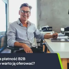 Uśmiechnięty mężczyzna w okularach siedzi przy biurku z laptopem, pisząc coś na papierze.