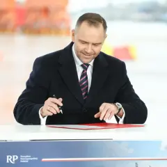 Mężczyzna w garniturze i płaszczu podpisuje dokumenty przy stole.