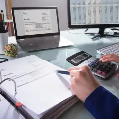 Osoba liczy faktury na kalkulatorze, analizując mechanizm podzielonej płatności od jakiej kwoty. Na biurku laptop, monitor z arkuszem kalkulacyjnym i dokumenty.