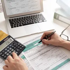 Dłoń pisząca na formularzu podatkowym PIT-36 obok kalkulatora i laptopa.