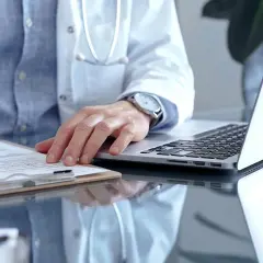 Lekarz w białym kitlu pisze na dokumentach, obok laptop.