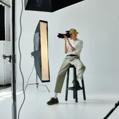 Fotograf siedzi na stołku w studiu, trzymając aparat i robiąc zdjęcie.
