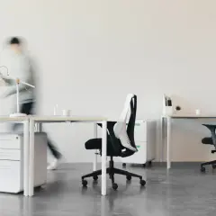 Nowoczesne biuro z białymi biurkami, krzesłami i szafkami. W tle widać poruszającą się postać, sugerującą dynamikę pracy i **firmantwo**.