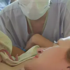 Matka z noworodkiem na piersi, w tle osoba w maseczce i fartuchu.