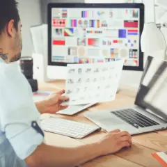 Mężczyzna przy biurku przegląda wydruki, przed nim komputer z kolorowymi grafikami i laptop.