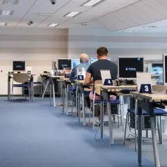 Rząd stanowisk komputerowych z osobami pracującymi, niebieska wykładzina.