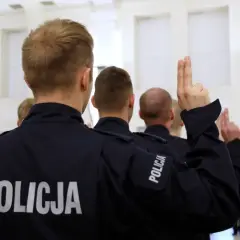Policjanci w mundurach z napisem "POLICJA" podnoszą ręce z dwoma palcami uniesionymi do góry.