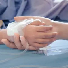 Dłoń dorosłego obejmuje dłoń dziecka z wenflonem. W tle pluszowy miś. Obraz symbolizuje wsparcie i troskę, jakiej wymaga zasilek pielegnacyjny.