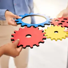 Dłonie łączą kolorowe trybiki, symbolizując współpracę i mechanizm.
