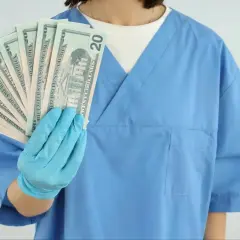 Higienistka stomatologiczna w niebieskim fartuchu trzyma wachlarz banknotów 20-dolarowych.