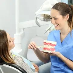 Uśmiechnięta higienistka stomatologiczna pokazuje pacjentce model szczęki i szczoteczkę.