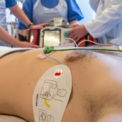 Pacjent z elektrodami EKG i defibrylatorem w tle, otoczony przez personel medyczny.
