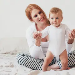 Uśmiechnięta mama z rudymi włosami pomaga 8-miesięcznemu dziecku w białym body wstać na łóżku.