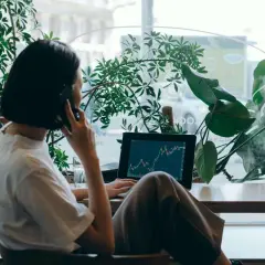 Kobieta rozmawia przez telefon przy laptopie z wykresem giełdowym, otoczona roślinami.