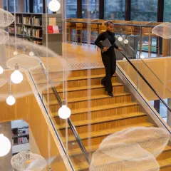 Kobieta z laptopem schodzi po drewnianych schodach w nowoczesnej bibliotece z ozdobnymi chmurkami i lampami.