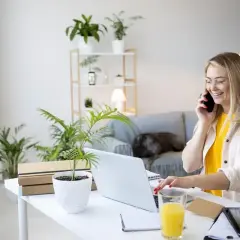 Uśmiechnięta kobieta pracuje przy laptopie i rozmawia przez telefon w domowym biurze.