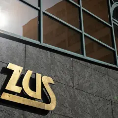 Złote logo ZUS na ciemnej, kamiennej elewacji budynku, nad nim okna z odbiciem.