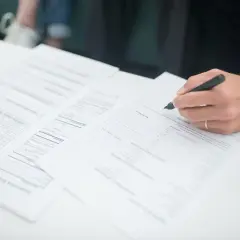 Dłoń z długopisem podpisująca dokumenty na białym stole.