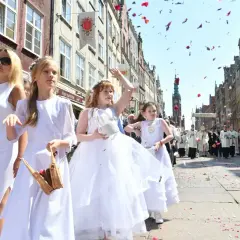 Procesja Bożego Ciała na ulicy, dzieci w białych strojach sypią płatki róż.