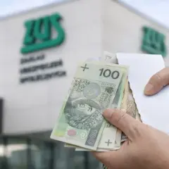 Dłonie trzymające polskie banknoty 100 zł przed budynkiem ZUS.
