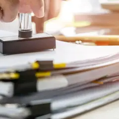 Dłoń trzymająca pieczątkę nad stosem dokumentów.