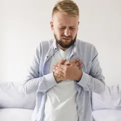 Mężczyzna trzymający się za klatkę piersiową z zamkniętymi oczami i grymasem bólu.