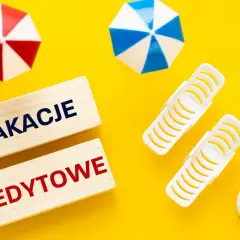 Drewniane klocki z napisem "Wakacje Kredytowe" na żółtym tle, otoczone miniaturowymi parasolkami plażowymi i kołami ratunkowymi.