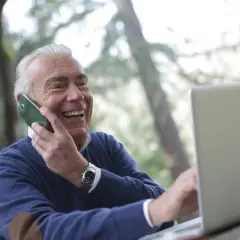 Uśmiechnięty starszy mężczyzna rozmawia przez telefon i pracuje na laptopie na zewnątrz.