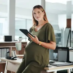 Ciężarna kobieta w zielonej sukience trzyma tablet i kubek w biurze.