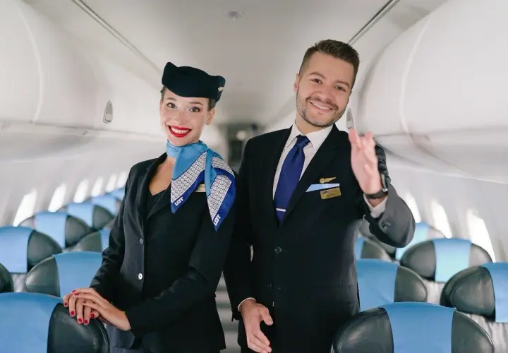 Uśmiechnięta stewardessa i steward w mundurach LOT stoją w samolocie, na tle rzędów siedzeń.