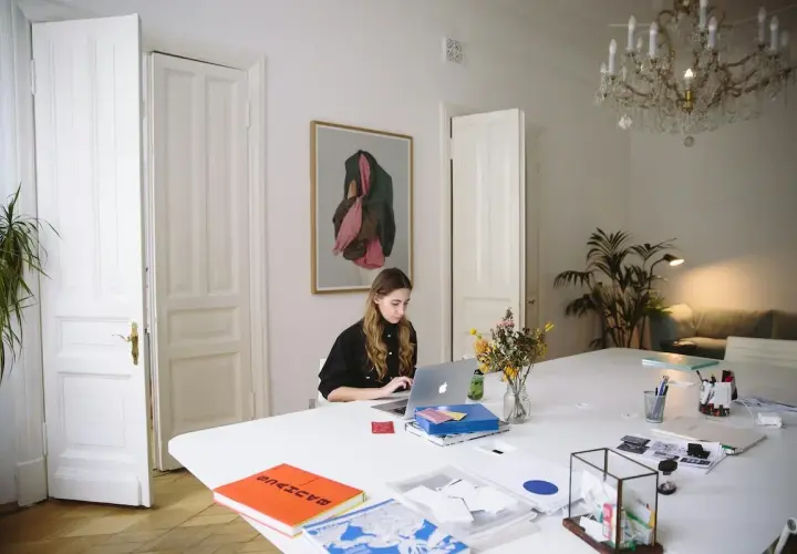 Kobieta pracuje przy laptopie w jasnym pomieszczeniu z obrazem na ścianie i bukietem kwiatów na stole.