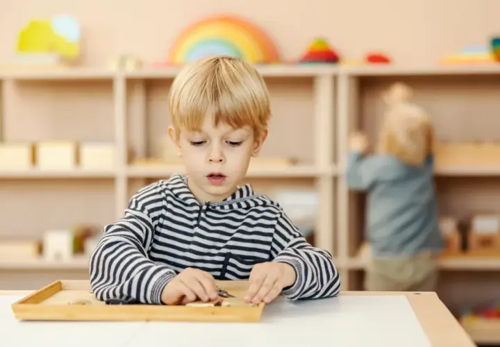 Chłopiec w bluzie w paski skupiony na zadaniu z materiałami Montessori. W tle inne dziecko przy półce.